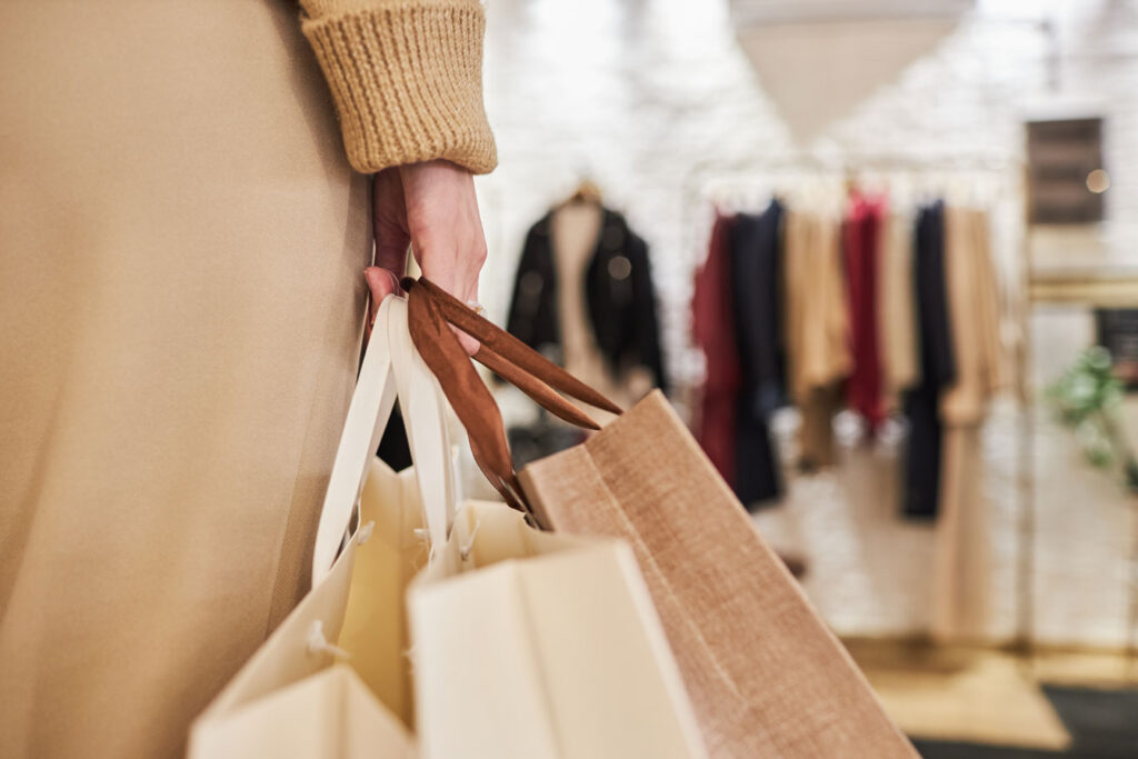 Close up view of a woman's had holding several shopping bags, in the distance are new clothing on hangers